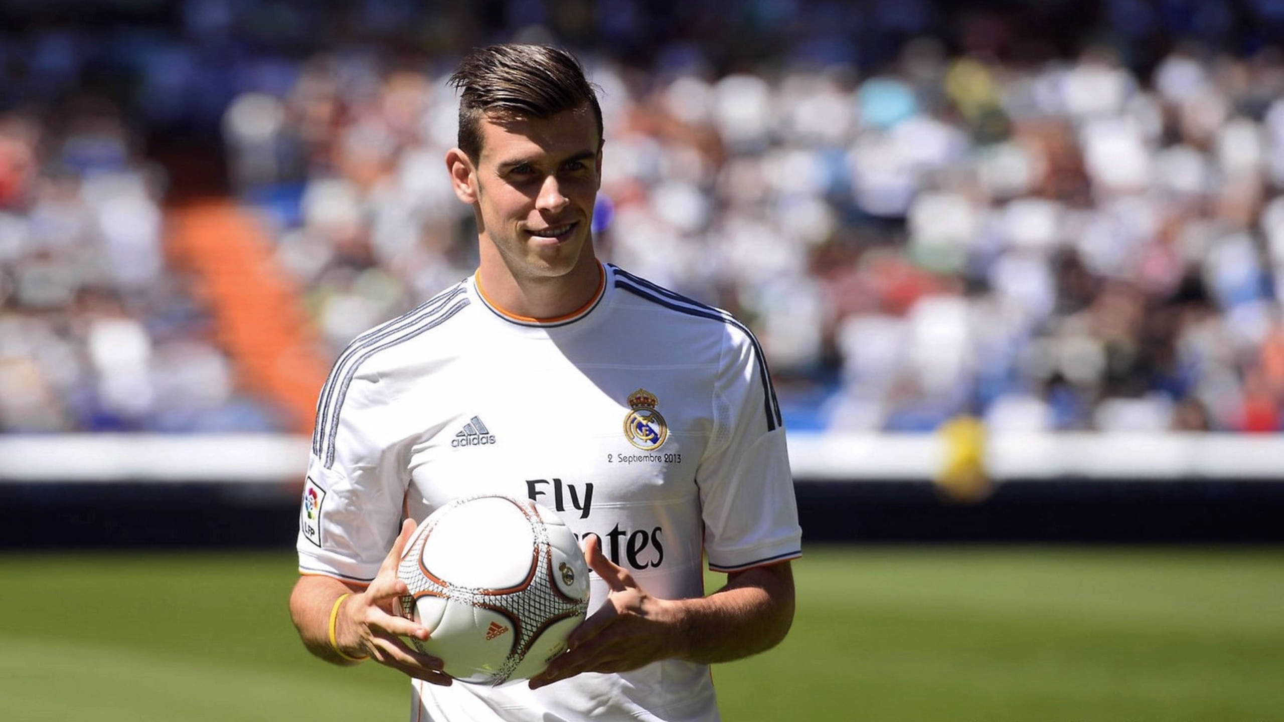 Gareth Bale 桌布 壁紙 — 一位微笑的足球員身穿白色皇家馬德里球衣，在陽光明媚的球場上抱著足球，背景是模糊的觀眾。