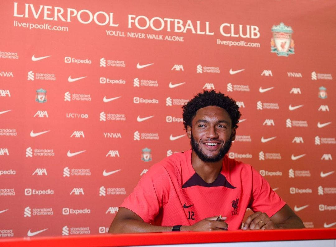 足球 桌布 壁紙 — 一位微笑的足球員身穿紅色利物浦球衣，坐在記者會桌前，背景為品牌紅色背景。