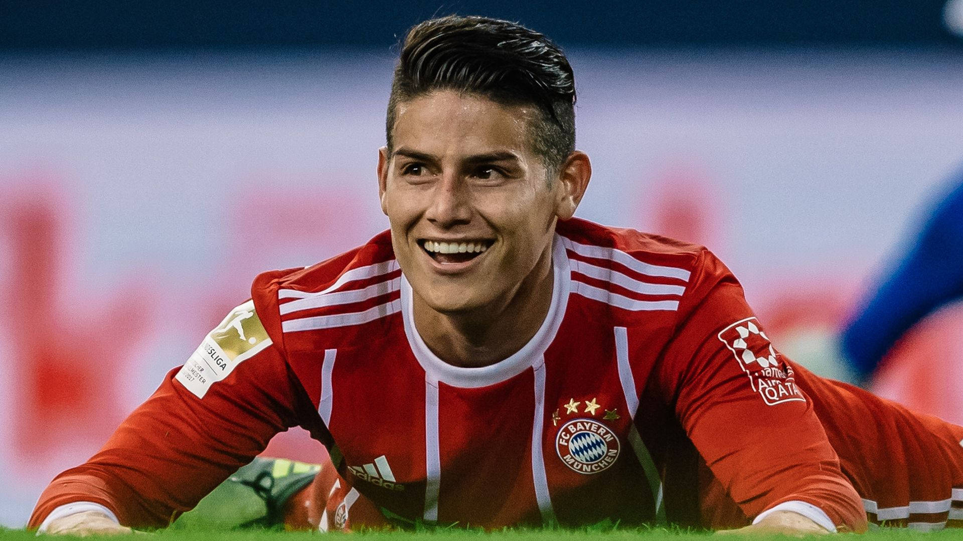 James Rodriguez 桌布 壁紙 — 一位微笑的足球員身穿紅色拜仁慕尼黑球衣，在球場燈光下比賽後躺在草地上。