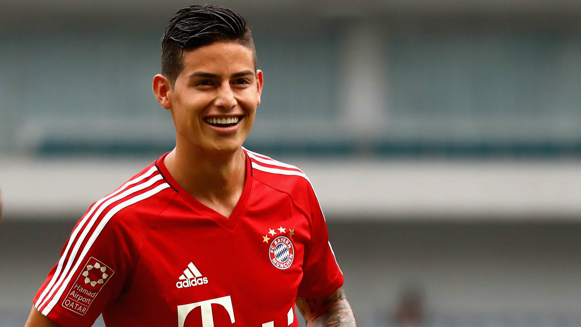James Rodriguez 桌布 壁紙 — 一位微笑的足球員身穿紅色拜仁慕尼黑球衣，站在球場上，身後是模糊的看台。