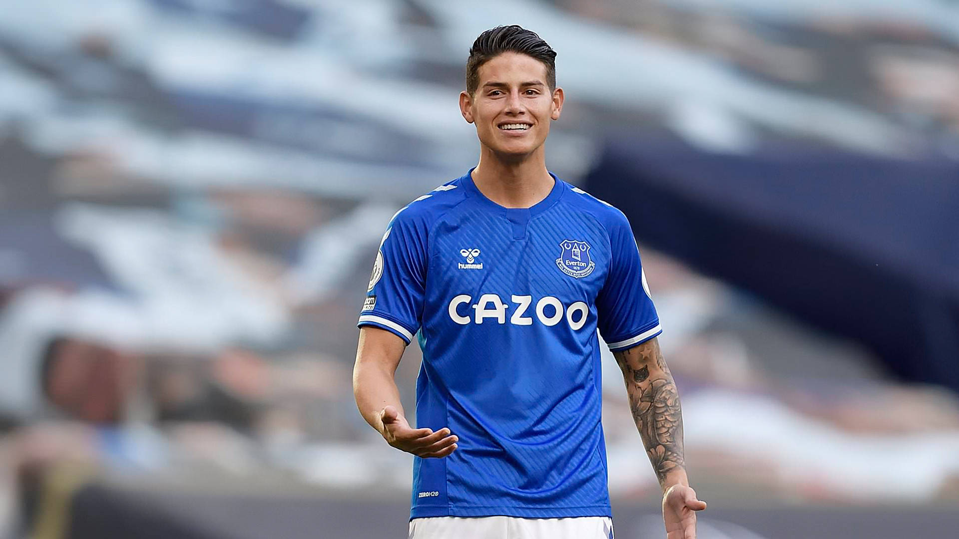 James Rodriguez 桌布 壁紙 — 一位微笑的足球員身穿藍色艾佛頓球衣，印有 'CAZOO' 贊助商標誌，站在球場上，背後是模糊的看台。