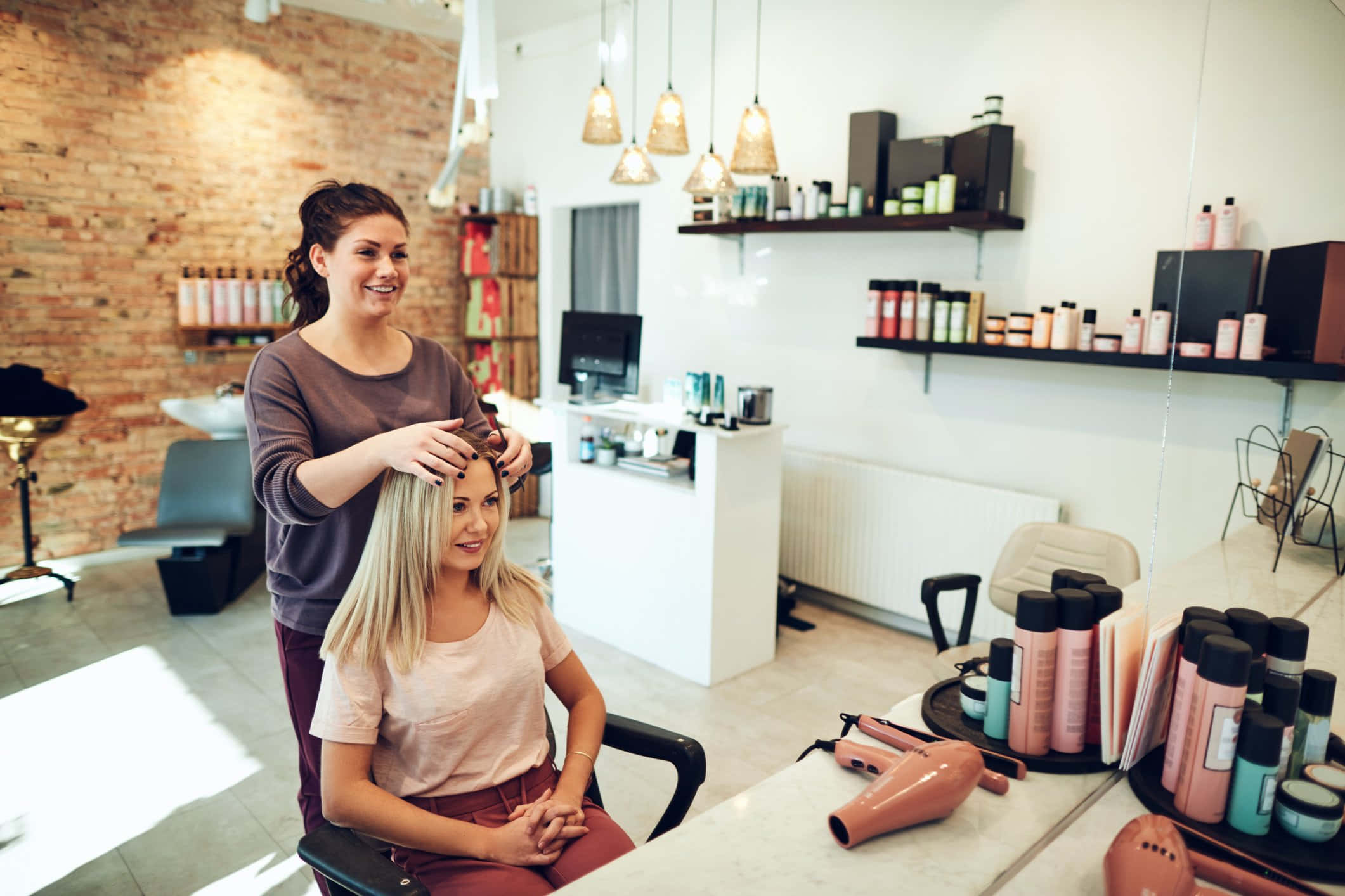 Hair Salon 桌布 壁紙 — 一位微笑的造型師在明亮現代、帶有裸露磚牆和吊燈的沙龍中，輕柔地為一位金髮客戶造型頭髮。