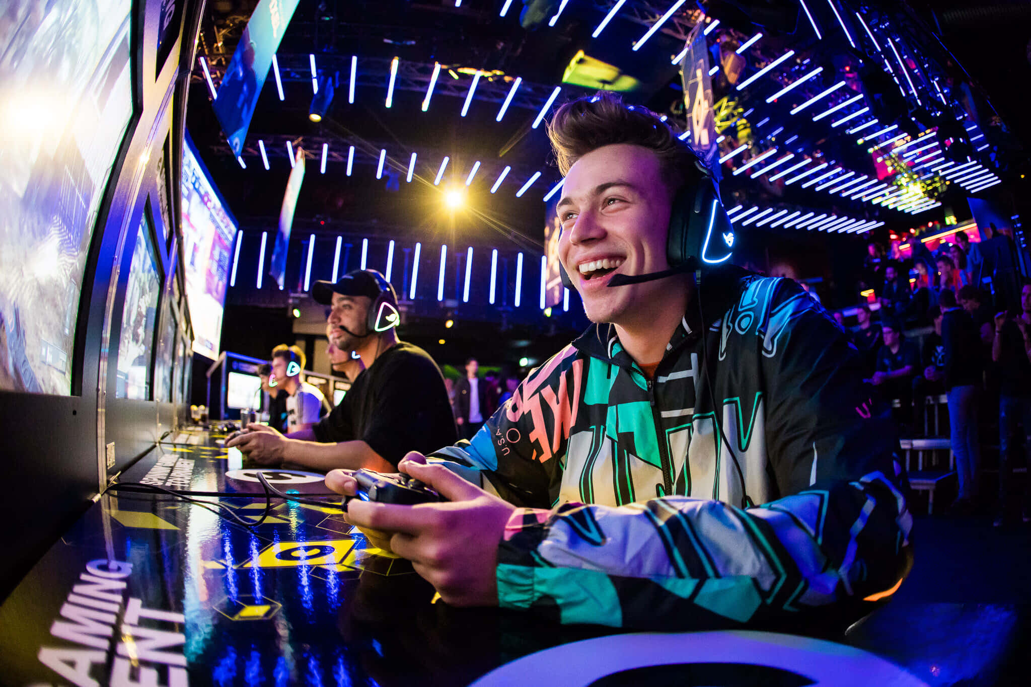 Gaming Tournaments 桌布 壁紙 — 一位微笑的遊戲玩家在充滿活力的電競場館中，在霓虹燈下拿著控制器大笑。