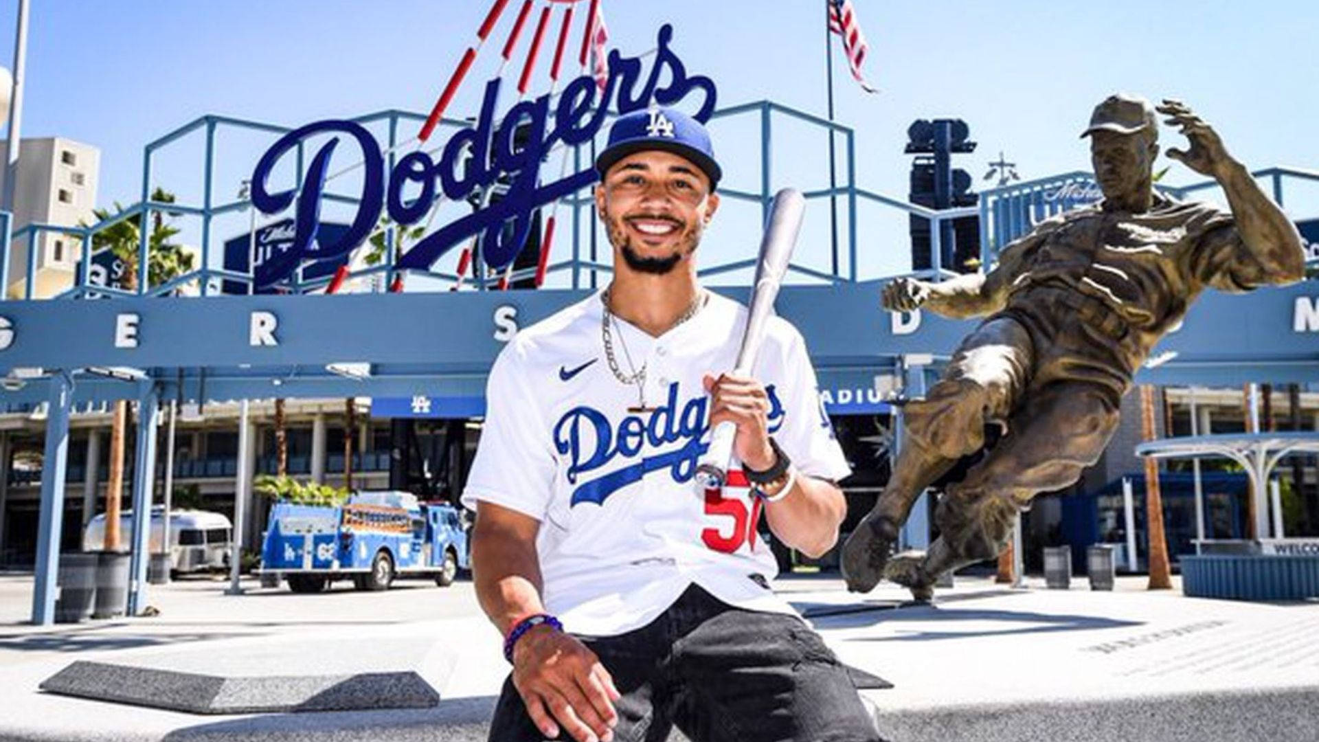 Mookie Betts 桌布 壁紙 — 一位微笑的道奇隊棒球運動員穿著制服，在標誌性的體育場標誌和銅像前拿著球棒擺姿勢。