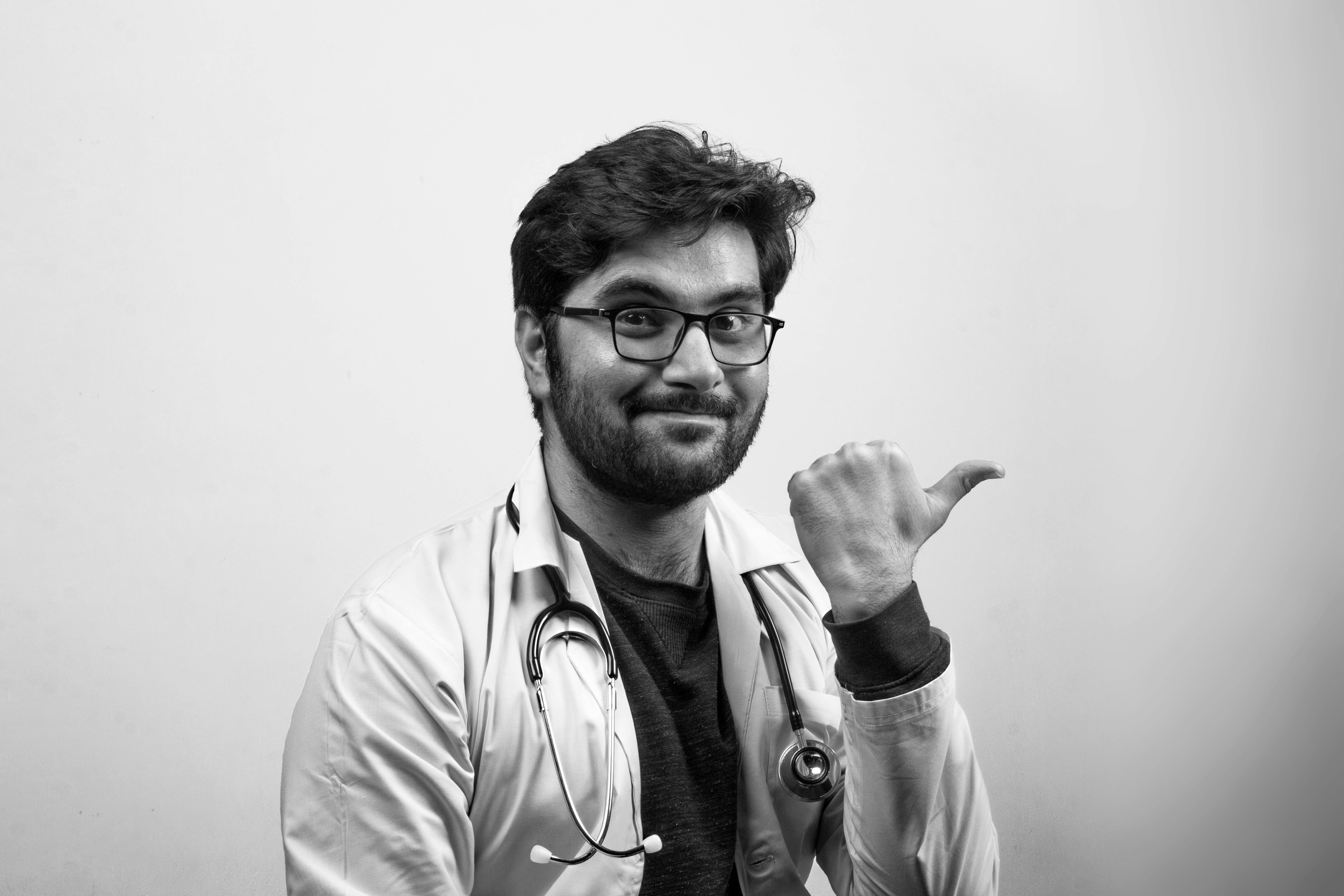 Doctors 桌布 壁紙 — 一位微笑的醫生穿著白袍和眼鏡，在純色背景下做出豎起大拇指的手勢。