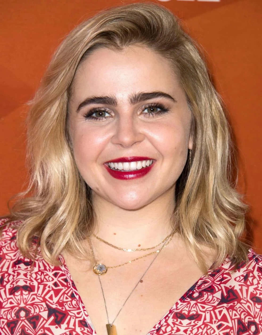 Mae Whitman 桌布 壁紙 — 一位微笑的金色波浪長髮、塗著大膽紅唇的女性，在溫暖的橙色背景前擺姿勢。