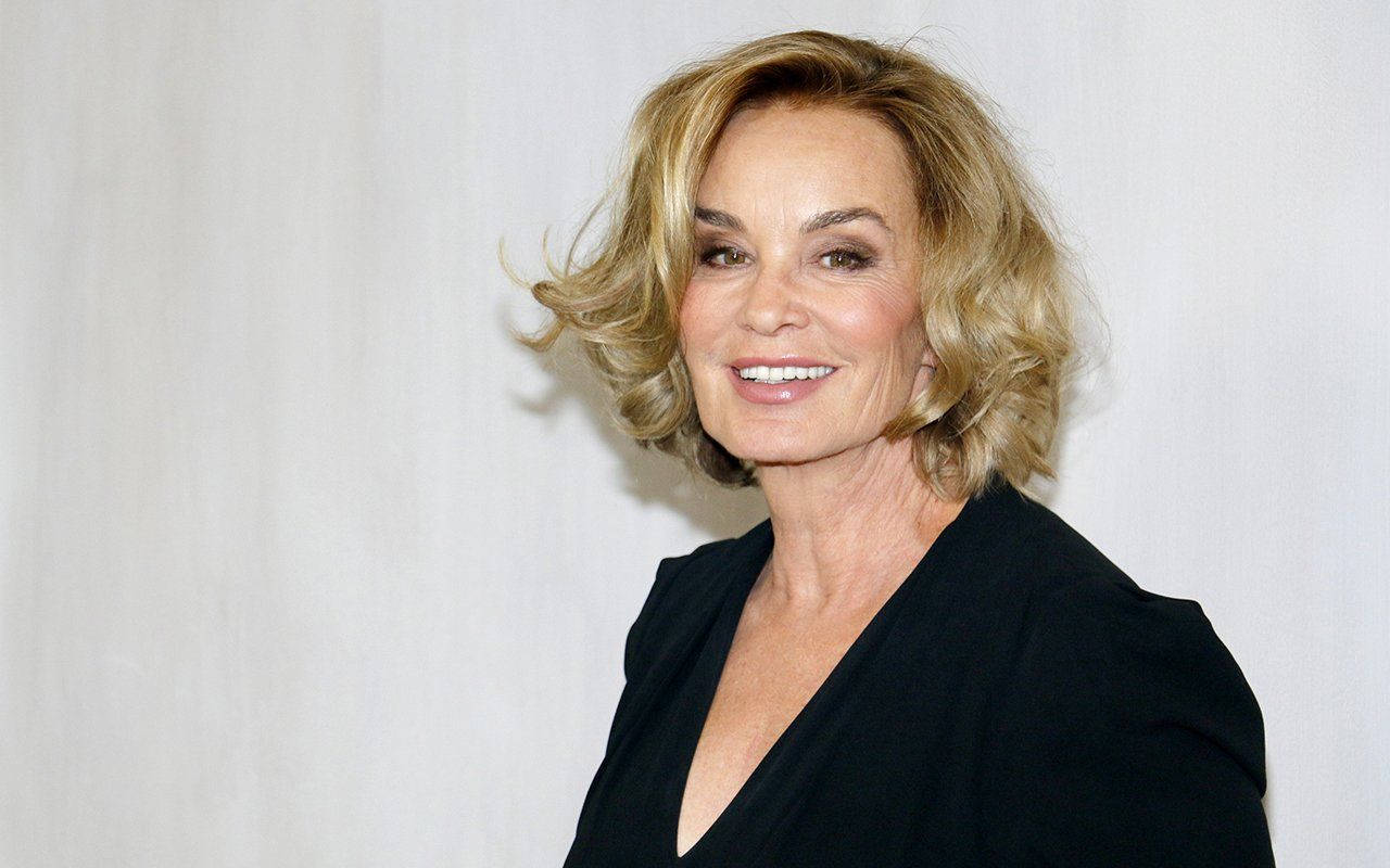 Jessica Lange 桌布 壁紙 — 一位微笑的金色波浪長髮女性，身穿黑色上衣，背景為純白色。