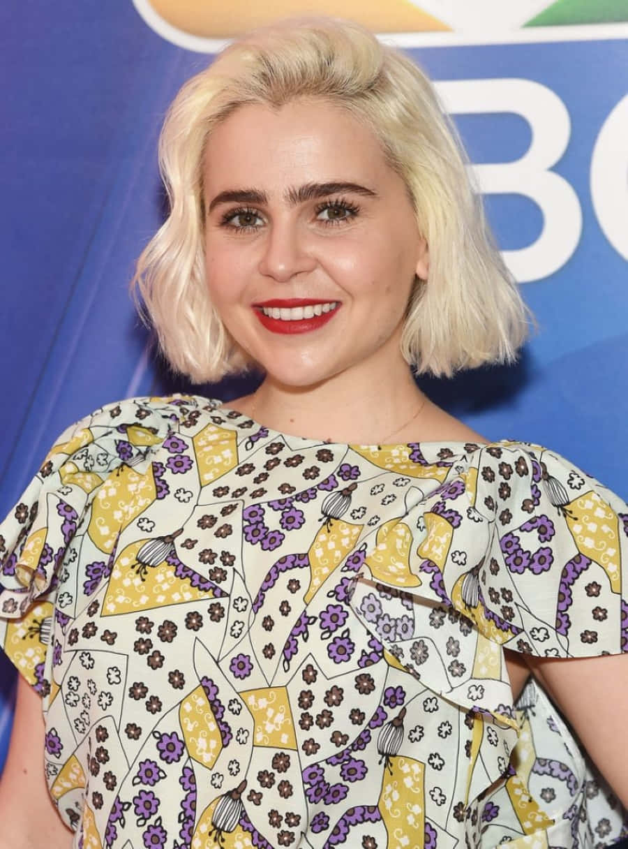 Mae Whitman 桌布 壁紙 — 一位微笑的金色鮑伯頭女性，塗著紅色唇膏，穿著黃紫色花卉圖案襯衫，背景為藍色品牌背景。