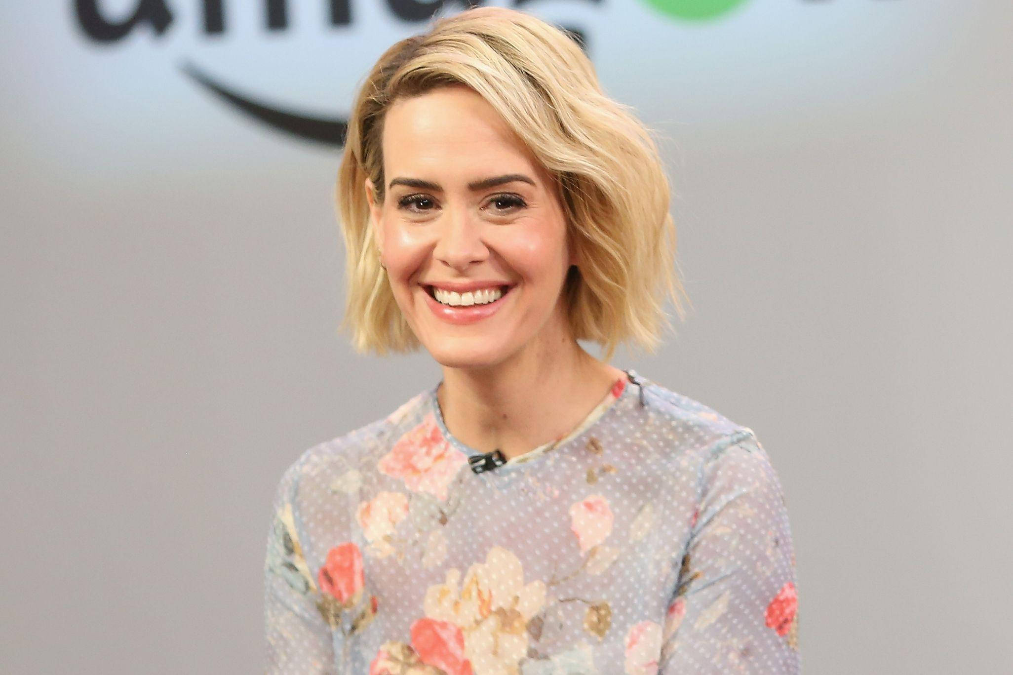 Celebrity 桌布 壁紙 — 一位微笑的金色鮑伯頭女性穿著花卉圖案上衣，背景是模糊的亞馬遜標誌。