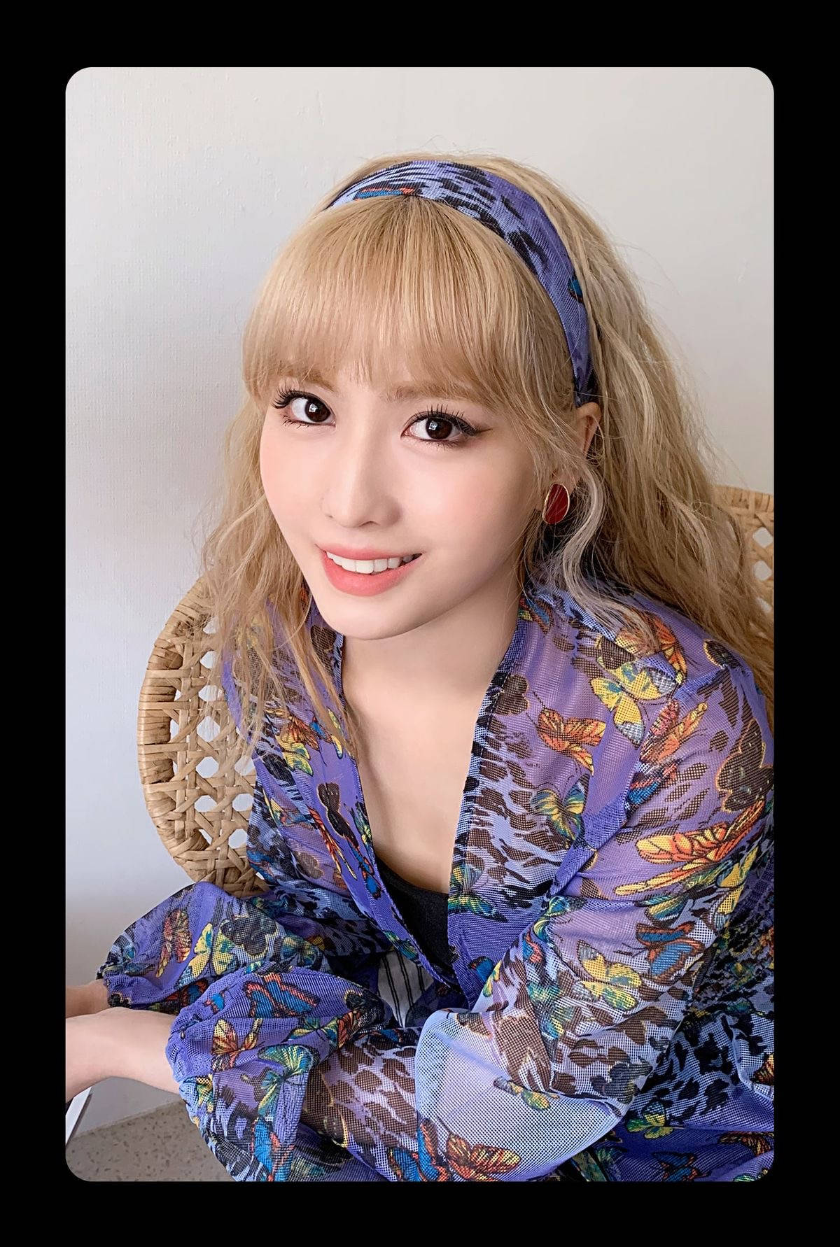 Momo 桌布 壁紙 — 一位微笑的金髮女子，身穿紫色蝴蝶圖案襯衫，坐在編織椅上，背景是素牆。