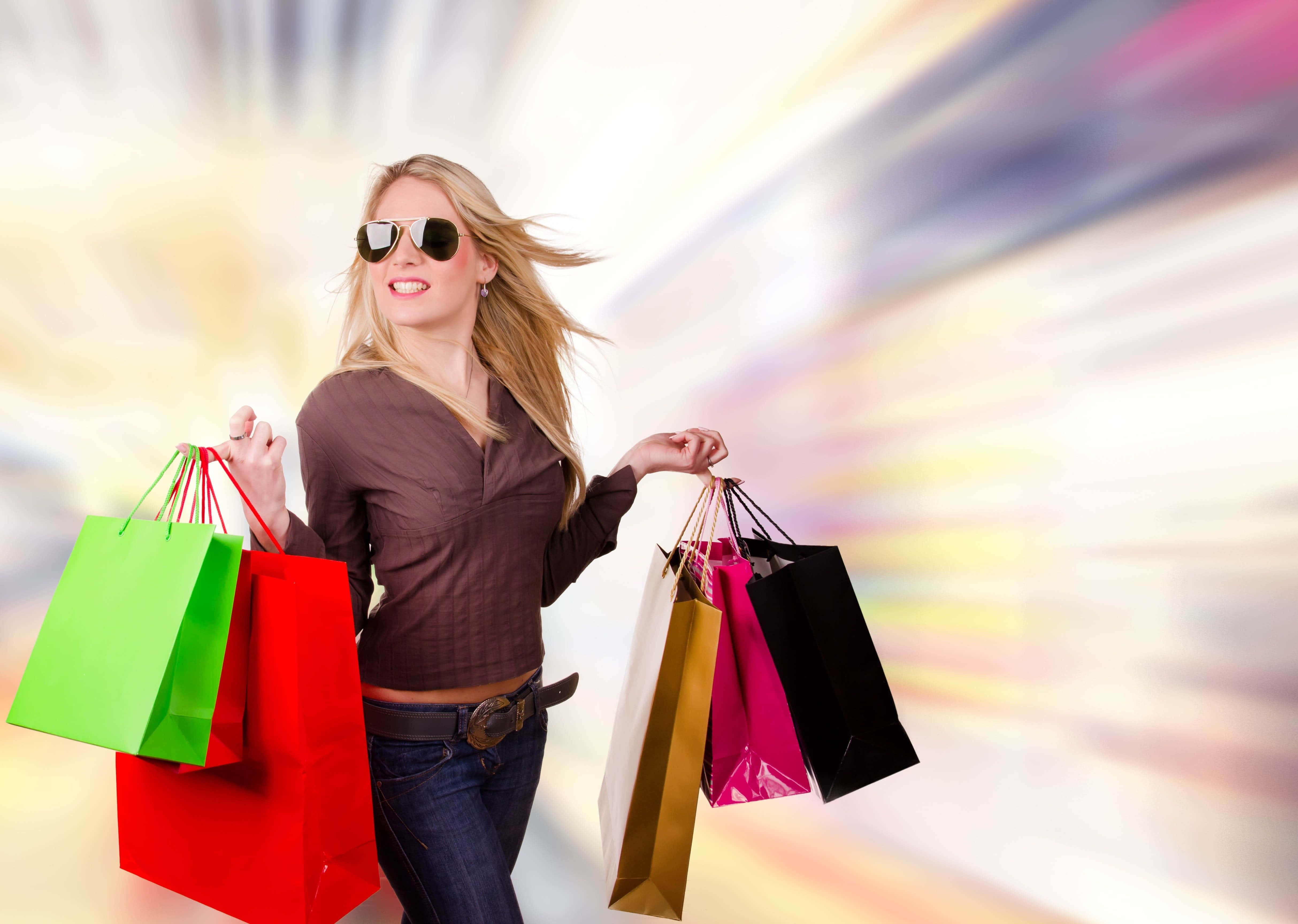 Shopping 桌布 壁紙 — 一位微笑的金髮女子戴著太陽眼鏡，在充滿動感的模糊背景下提著色彩繽紛的購物袋。