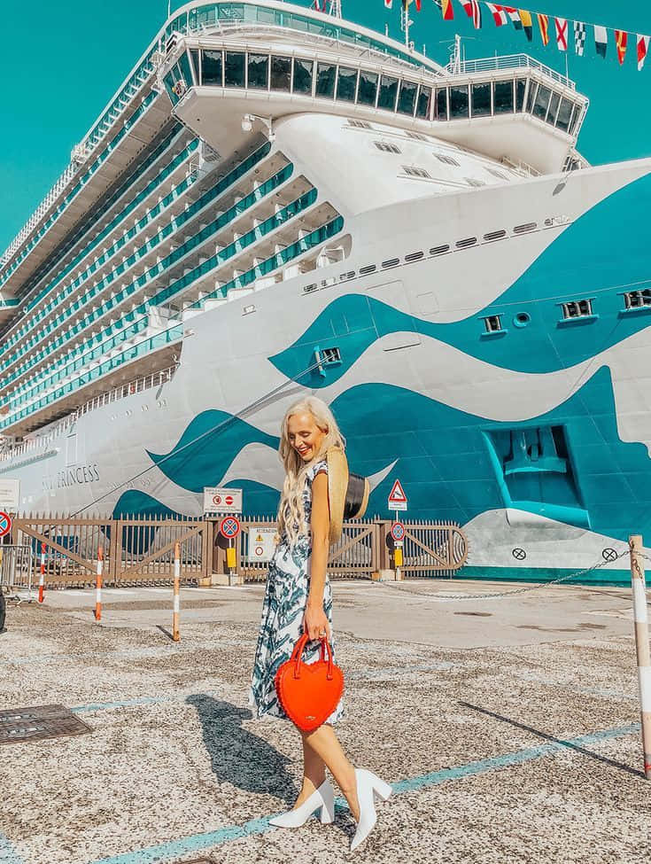 Cruise 桌布 壁紙 — 一位微笑的金髮女子穿著圖案連衣裙和白色高跟鞋，在晴朗的藍天下自信地走過一艘巨大的藍綠色和白色郵輪。