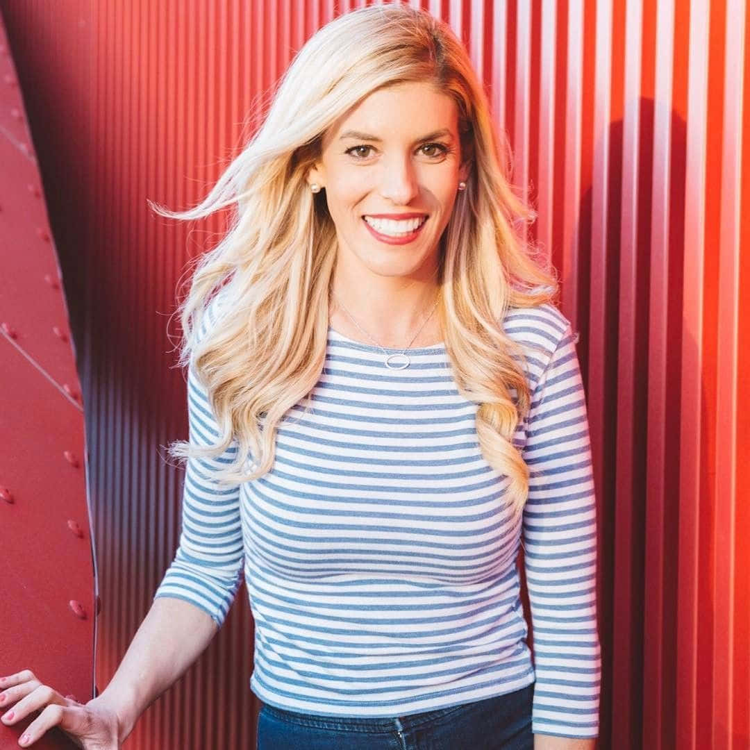 Rebecca Zamolo 桌布 壁紙 — 一位微笑的金髮女子穿著條紋襯衫，自信地站在鮮紅色的波紋金屬牆前。