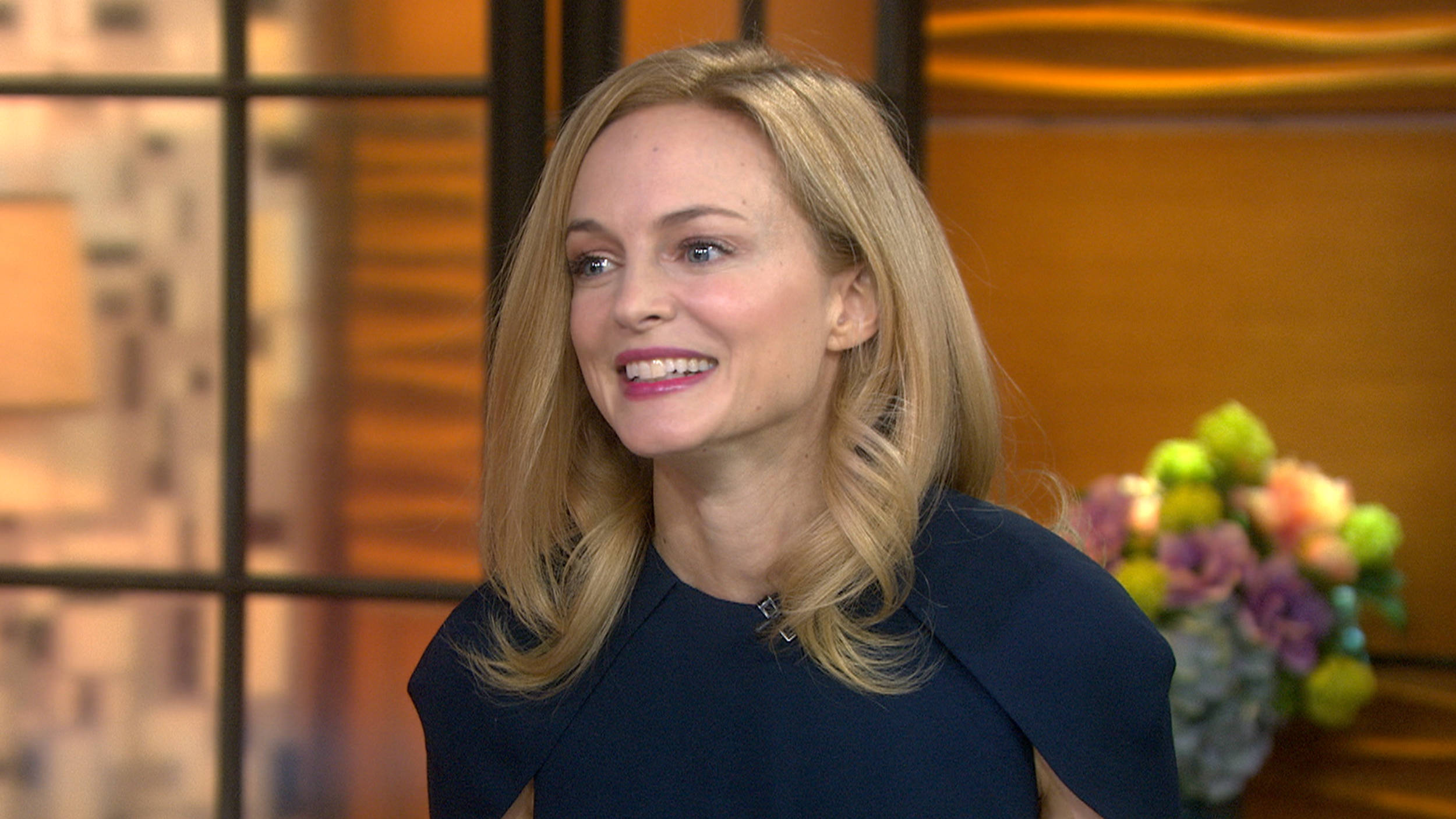 Heather Graham 桌布 壁紙 — 一位微笑的金髮女子穿著深藍色連衣裙，坐在溫暖照明的攝影棚中，身後是模糊的城市天際線和色彩繽紛的花朵。