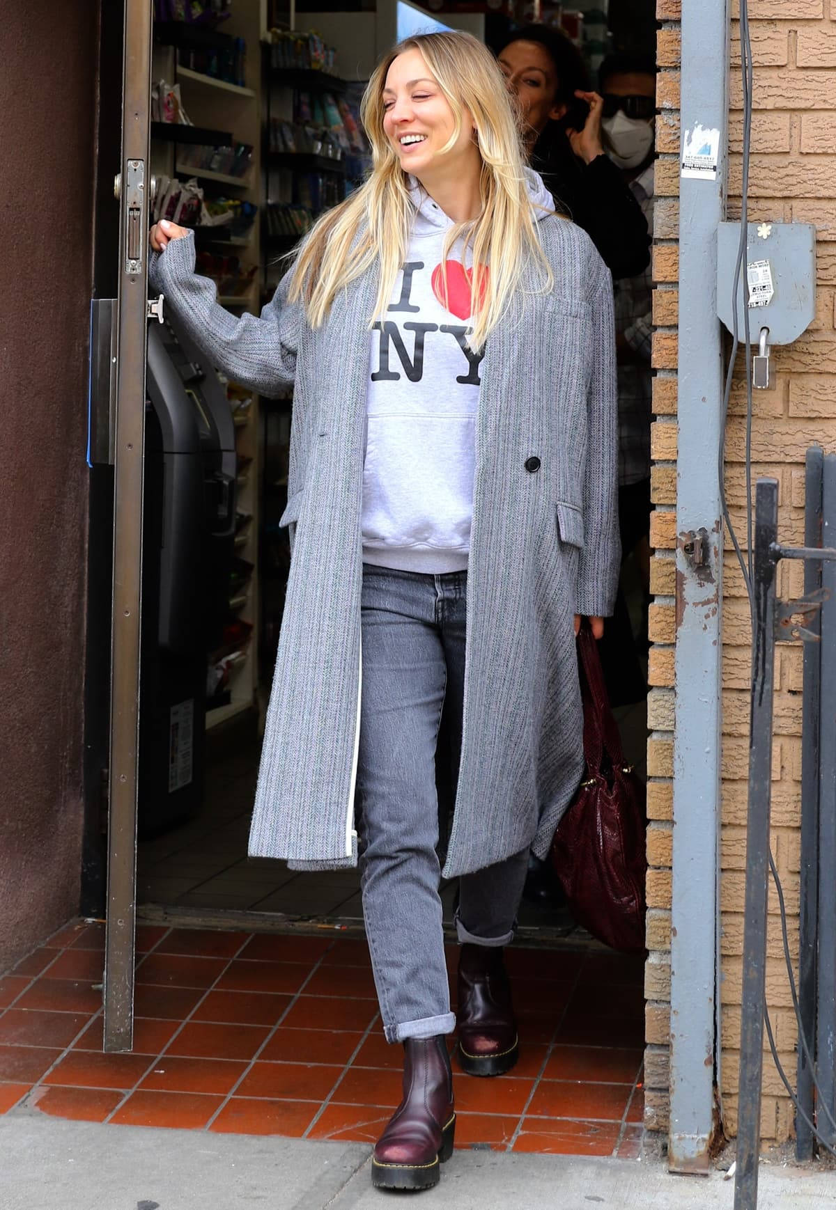 Kaley Cuoco 桌布 壁紙 — 一位微笑的金髮女子穿著灰色外套和I heart NY衛衣，從一棟紅磚地板建築物中走出。