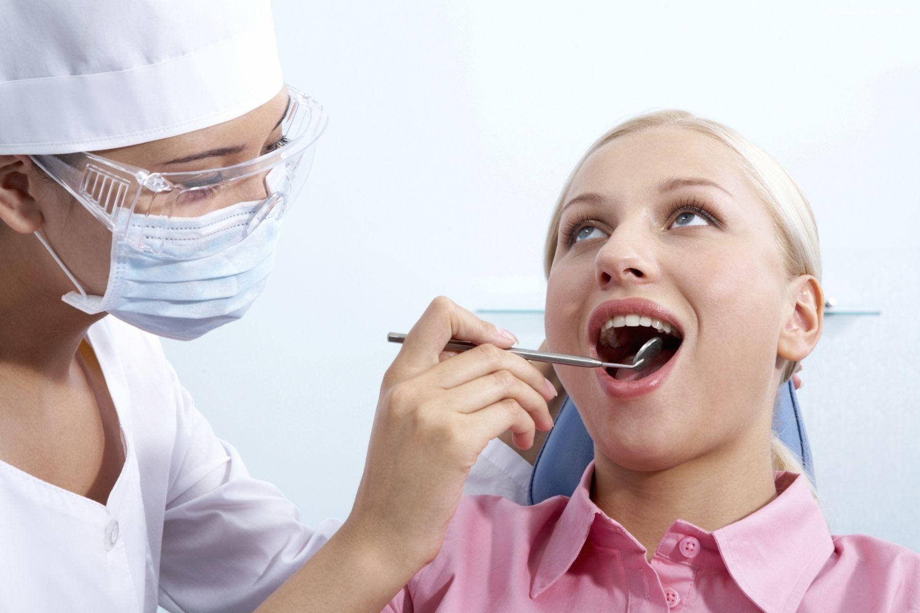 Dentistry 桌布 壁紙 — 一位微笑的金髮女子穿著粉紅色襯衫，張大嘴巴，一位戴著口罩的白袍牙醫用金屬工具檢查她的牙齒。