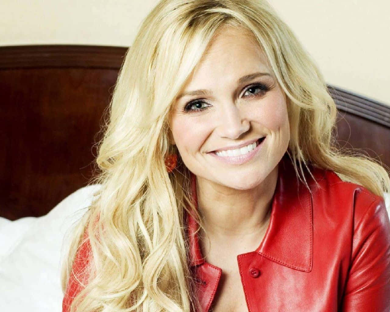 kristin chenoweth 桌布 壁紙 — 一位微笑的金髮女子穿著紅色皮夾克，坐在有木製床頭板的床上。