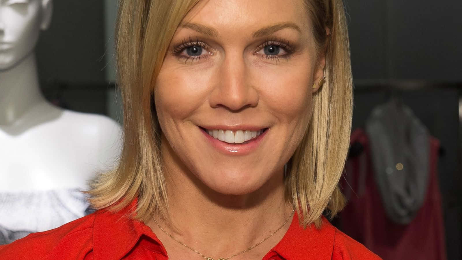 Jennie Garth 桌布 壁紙 — 一位微笑的金髮女子穿著紅色襯衫的特寫肖像，背景有模特兒和衣架。
