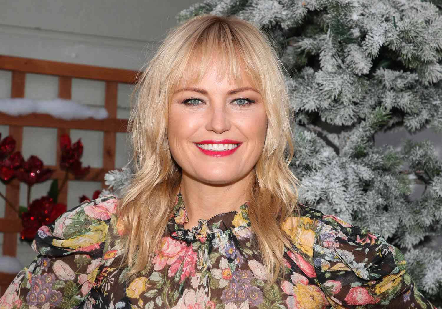 Actress HD 桌布 壁紙 — 一位微笑的金髮女子穿著花卉圖案襯衫，站在一棵覆蓋白雪的常青樹前，樹上掛著紅色聖誕裝飾。