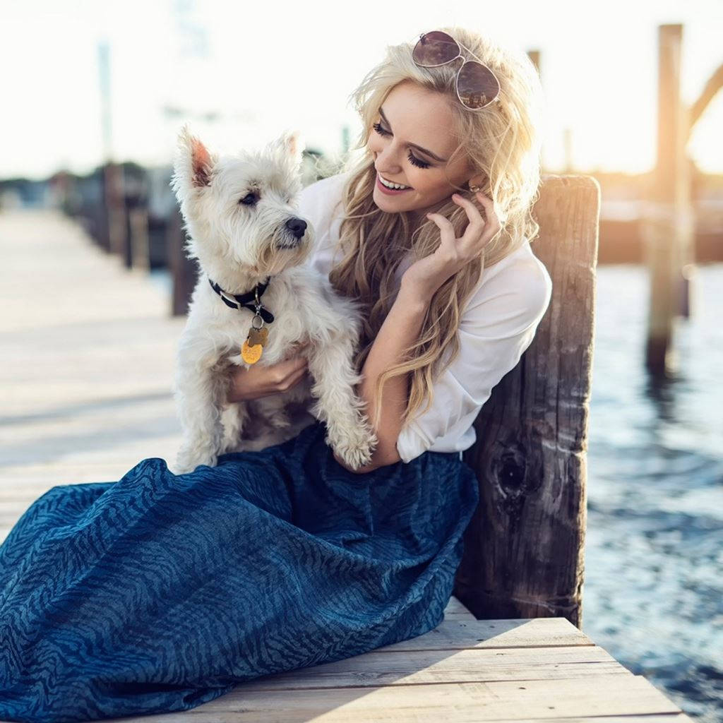 Dog And Girl 桌布 壁紙 — 一位微笑的金髮女子穿著藍色裙子，在黃金時段於木碼頭上抱著一隻白色的西部高地白㹴。