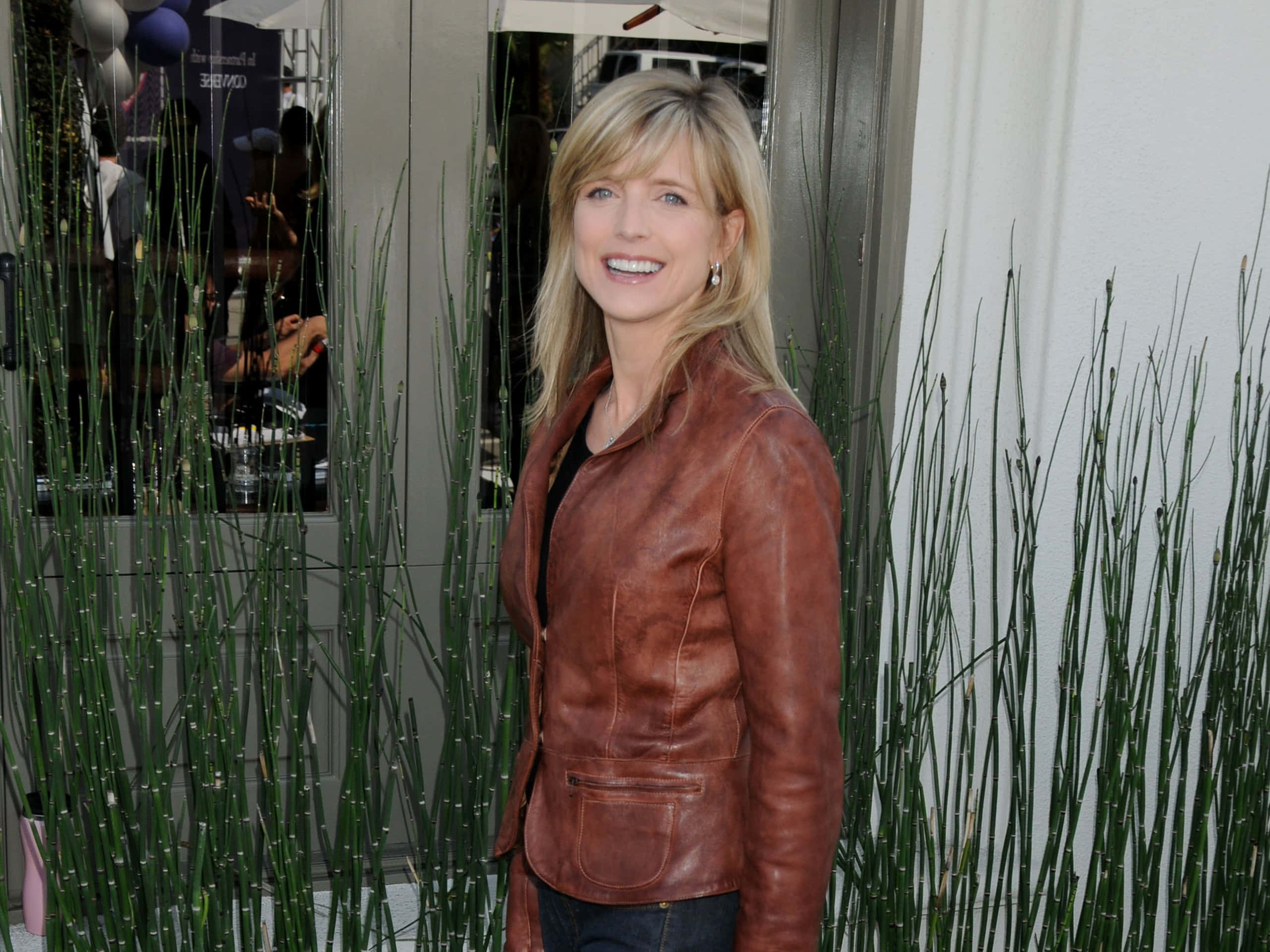 Courtney Thorne Smith 桌布 壁紙 — 一位微笑的金髮女子身穿棕色皮夾克，站在玻璃門附近高高的綠色蘆葦旁。