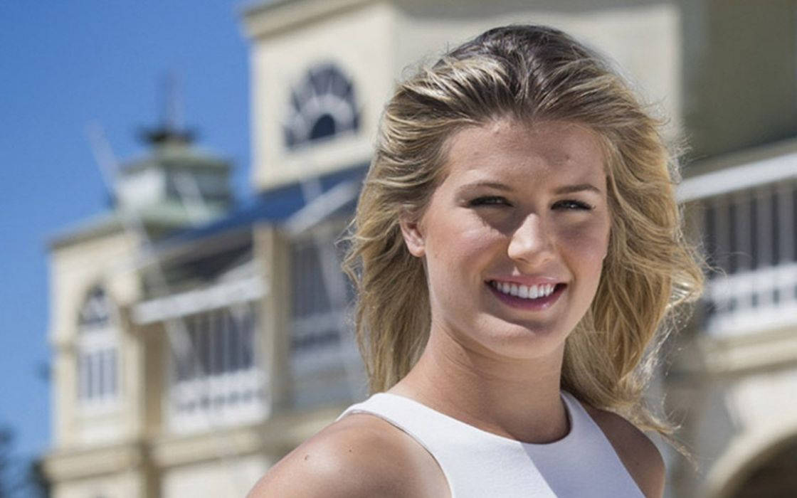 Eugenie Bouchard 桌布 壁紙 — 一位微笑的金髮女子身穿白色上衣，站在晴朗藍天下陽光普照的鐘樓前。