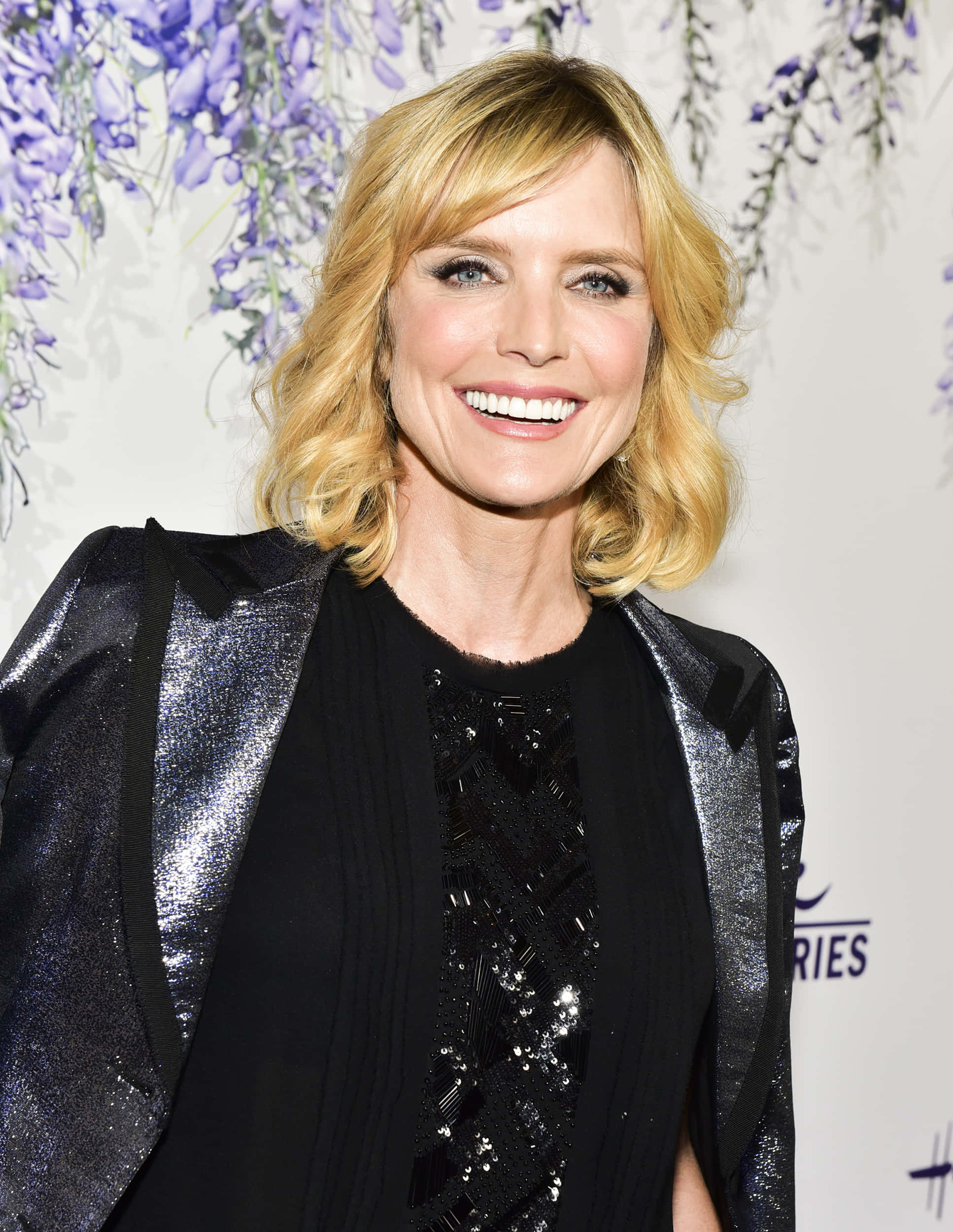 Courtney Thorne Smith 桌布 壁紙 — 一位微笑的金髮女子身穿黑色亮片服裝，站在紫色紫藤花背景前。