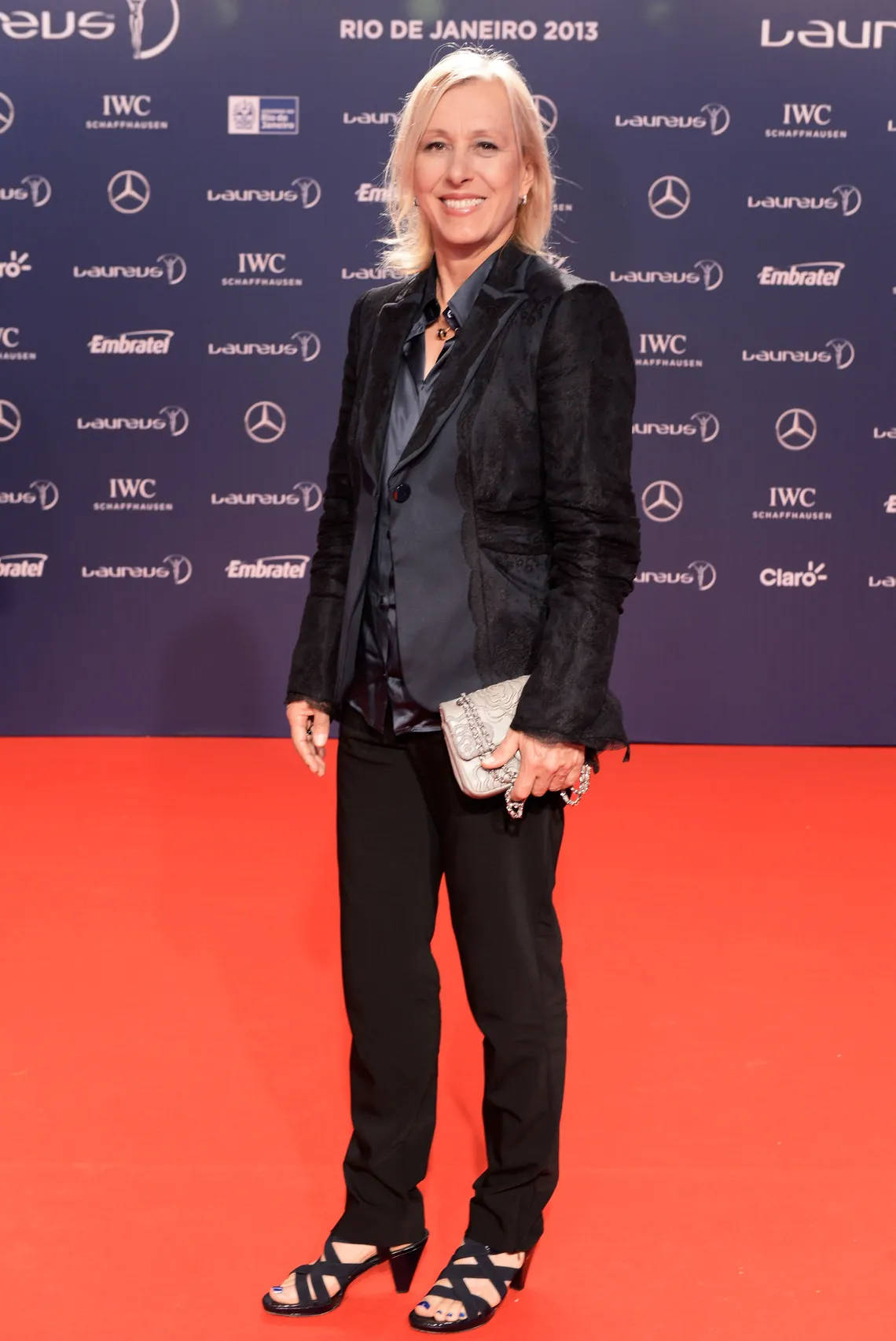 Martina Navratilova 桌布 壁紙 — 一位微笑的金髮女子身穿黑色西裝和高跟鞋，站在2013年里約熱內盧活動的紅地毯上，背景是帶有品牌的藍色牆壁。