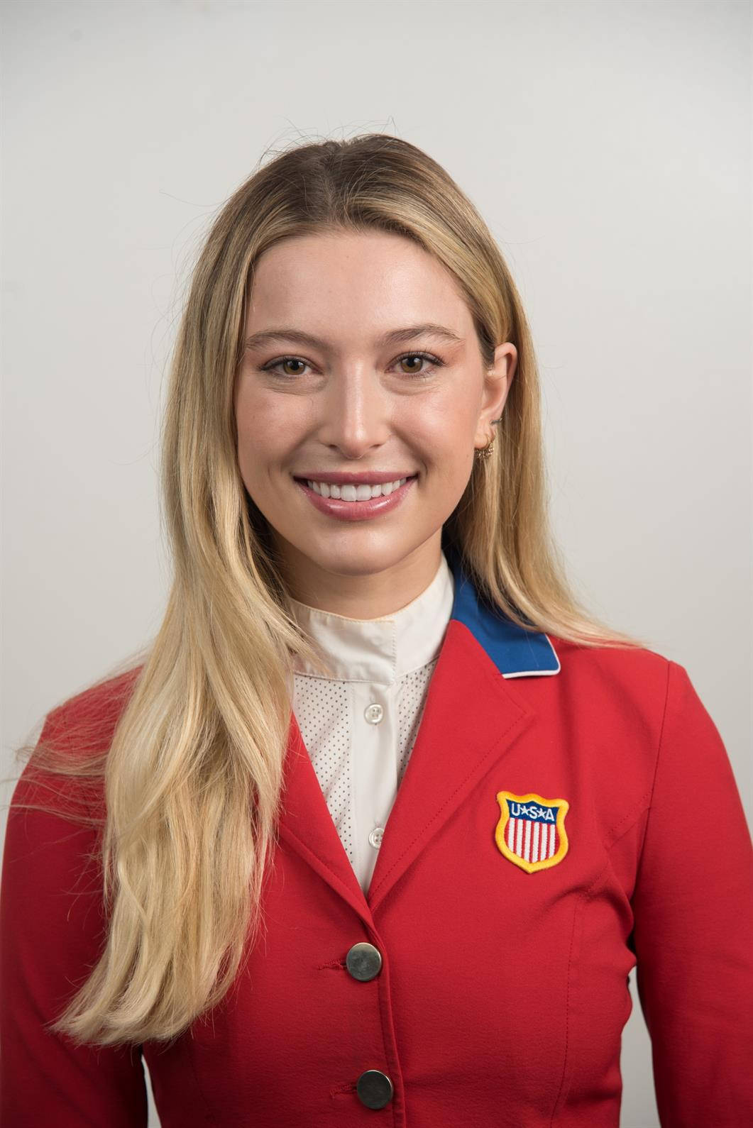 Athletic 桌布 壁紙 — 一位微笑的金髮女性，身穿紅色美國隊西裝外套，內搭白色有領襯衫，背景為純灰色。