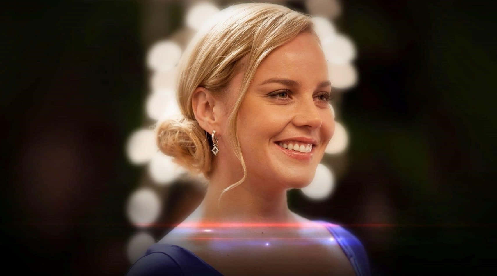 Abbie Cornish 桌布 壁紙 — 一位微笑的金髮女性，身穿藍色連衣裙，優雅的盤髮和鑽石耳環，背景為散景和鏡頭光暈。