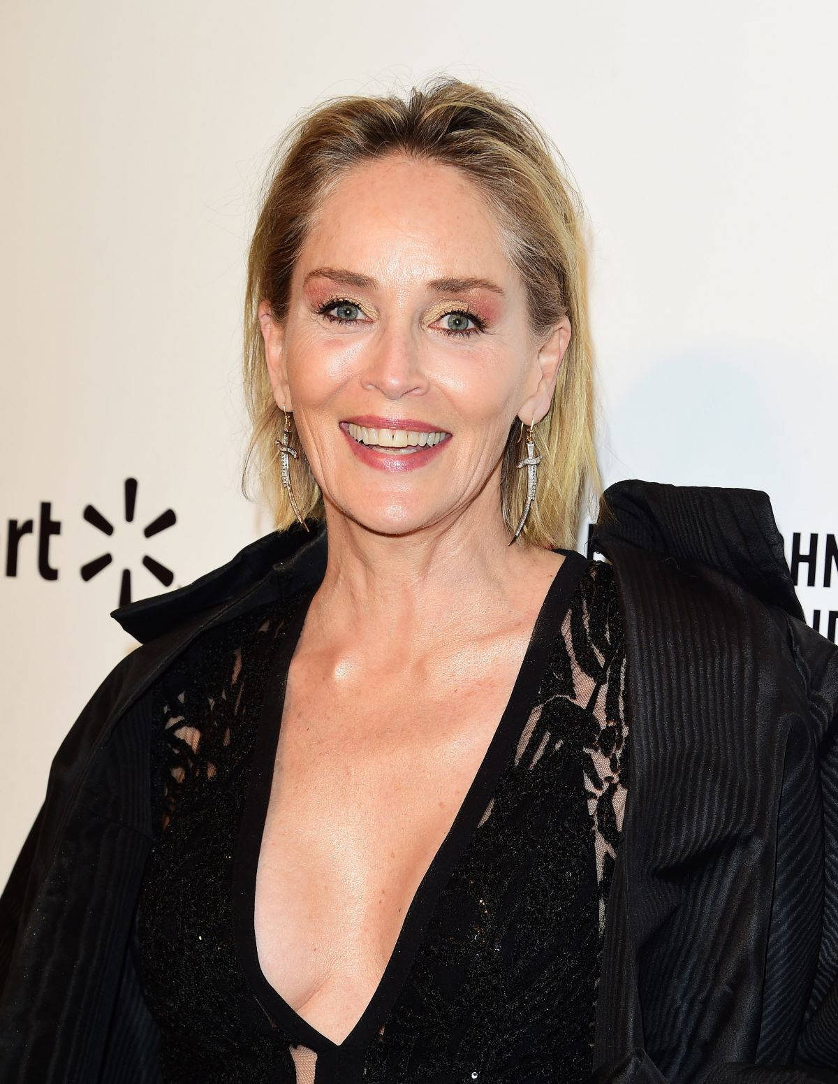 Celebrity 桌布 壁紙 — 一位微笑的金髮女性在帶有品牌背景的活動中穿著黑色蕾絲連衣裙和外套。