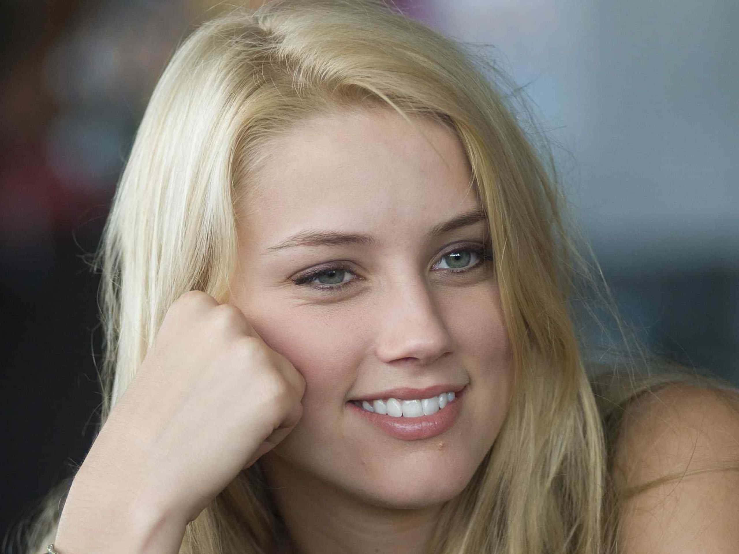 Amber Heard 桌布 壁紙 — 一位微笑的金髮女性在柔和自然光下將臉頰靠在拳頭上的特寫肖像。