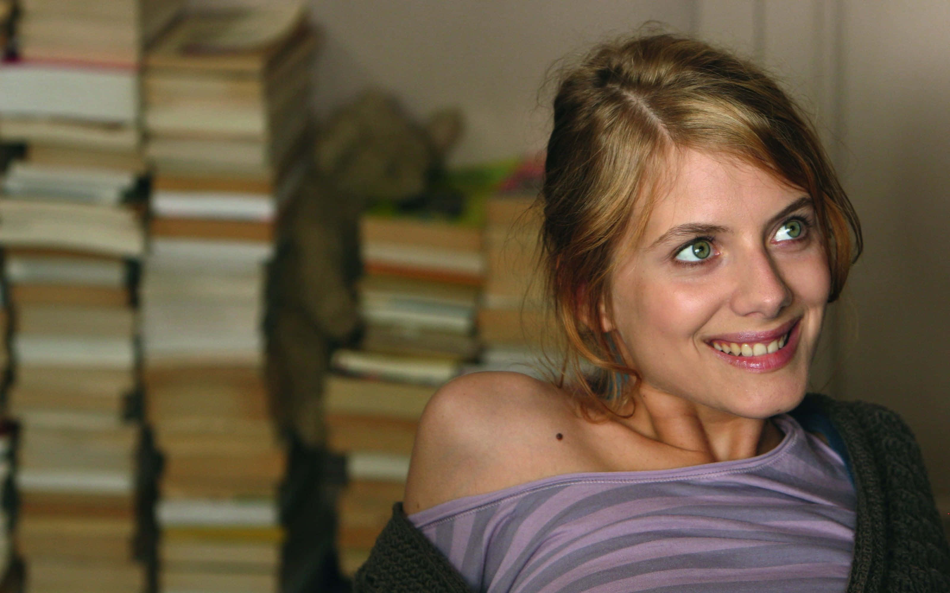 Mélanie Laurent 桌布 壁紙 — 一位微笑的金髮女性在舒適的室內環境中，旁邊是高聳的書堆，視線離開鏡頭。