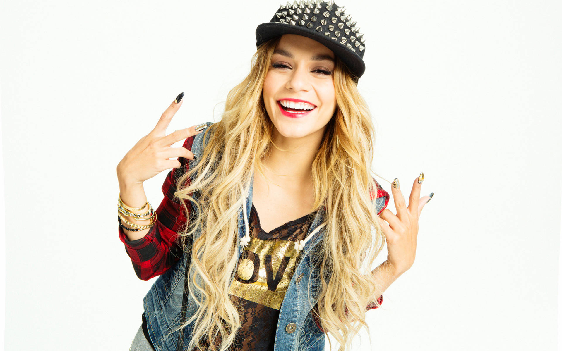 Vanessa Hudgens 桌布 壁紙 — 一位微笑的金髮女性戴著鉚釘帽和牛仔背心，在純白背景前做出搖滾手勢。