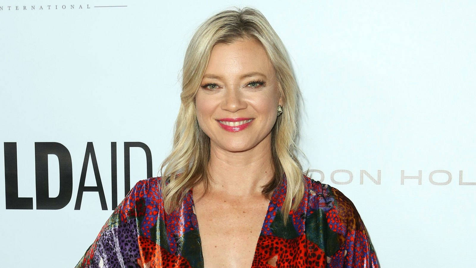 Celebrity 桌布 壁紙 — 一位微笑的金髮女性穿著彩色圖案上衣，在淺藍色背景前擺姿勢，背景帶有部分文字。