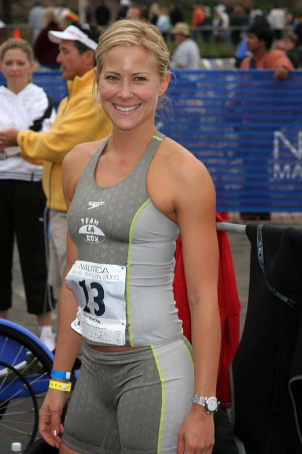 Brittany Daniel 桌布 壁紙 — 一位微笑的金髮女性穿著灰色鐵人三項服裝，帶有比賽號碼13，站在鐵人三項賽事中，身後有觀眾和藍色護欄。