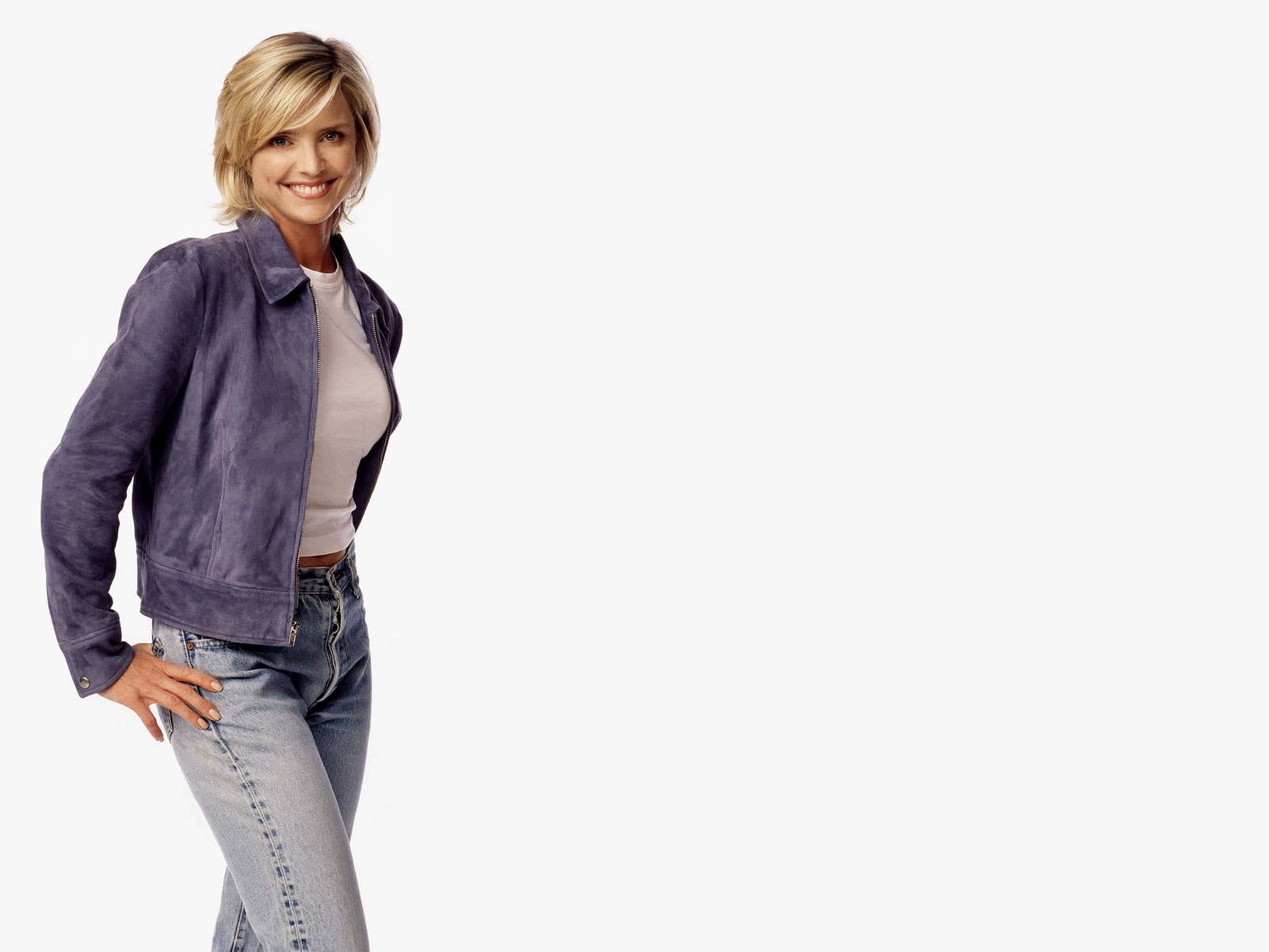 Courtney Thorne Smith 桌布 壁紙 — 一位微笑的金髮女性穿著紫色麂皮外套和牛仔褲，在純白背景前自信地擺姿勢。