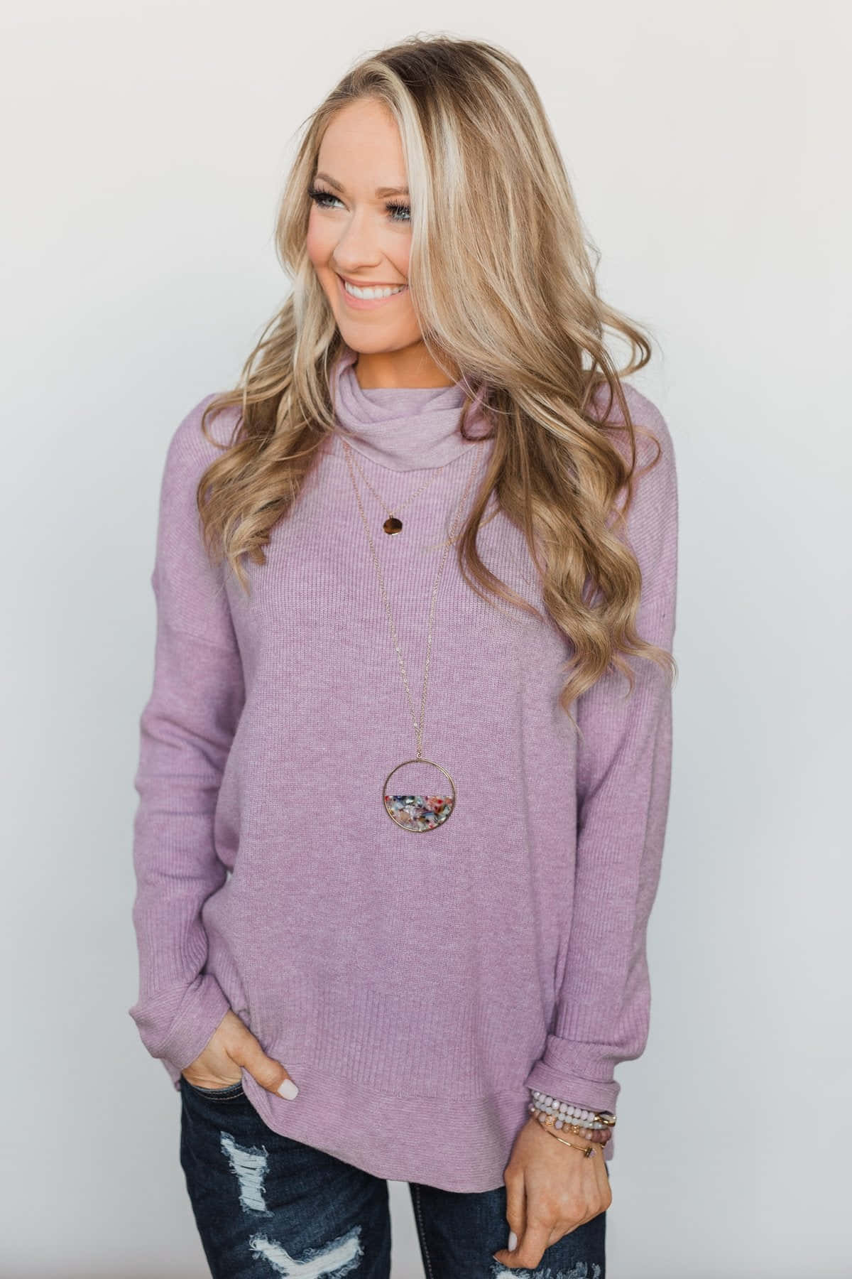 Purple Turtle-neck Sweater 桌布 壁紙 — 一位微笑的金髮女性穿著薰衣草色高領毛衣和破洞牛仔褲，在純白背景前擺姿勢。