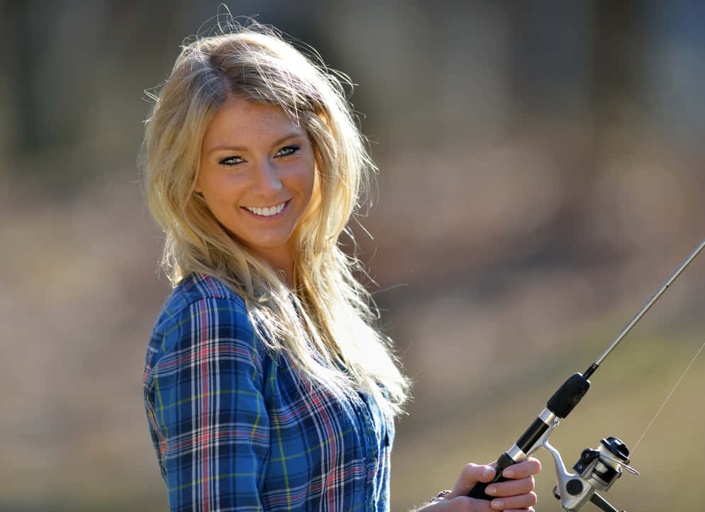 Country Girl 桌布 壁紙 — 一位微笑的金髮女性穿著藍色格紋襯衫，在柔和模糊的戶外背景下拿著釣魚竿。