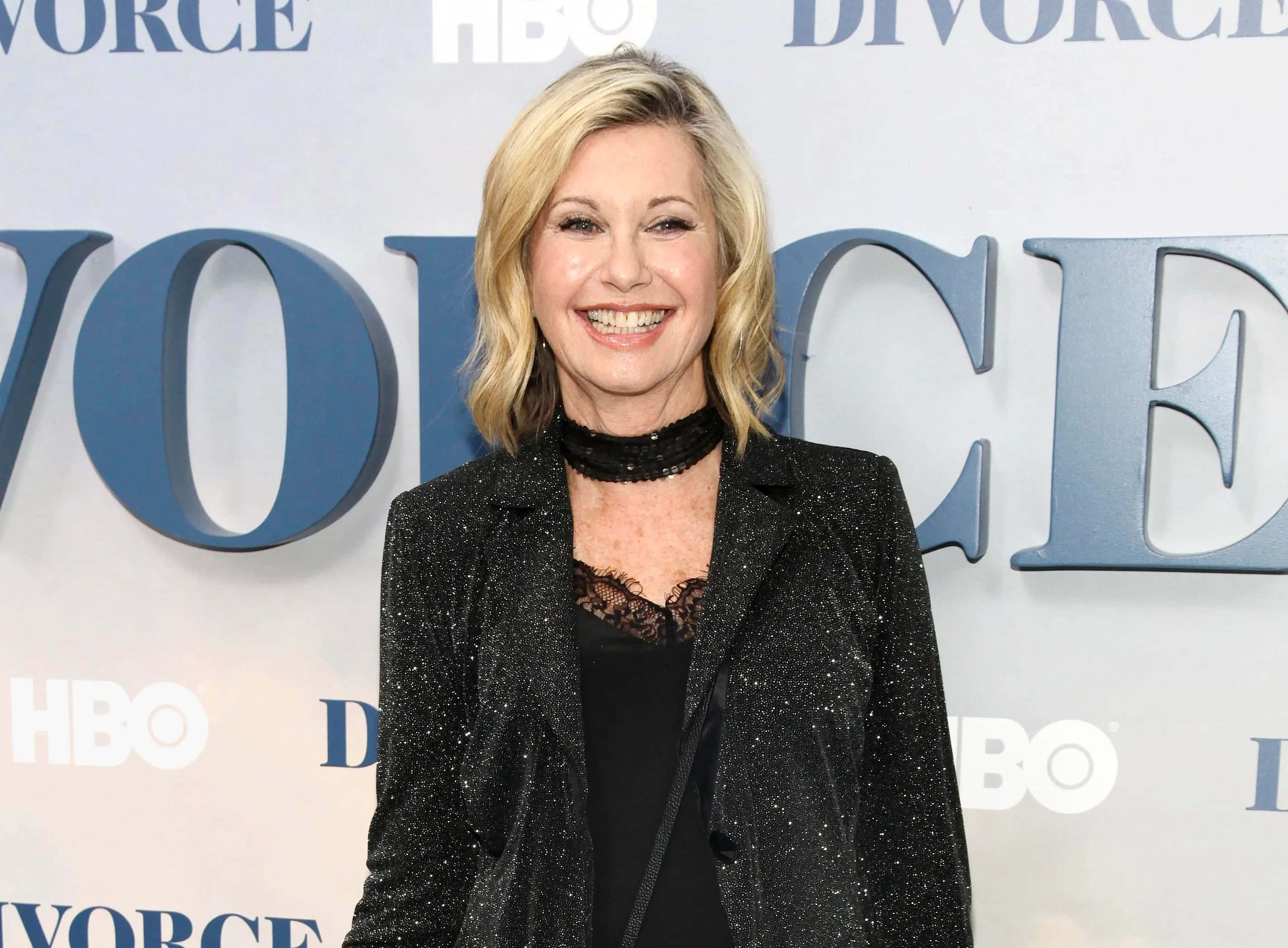 Olivia Newton John 桌布 壁紙 — 一位微笑的金髮女性穿著閃亮的黑色西裝外套，站在藍色HBO Divorce背景前，出席紅毯活動。