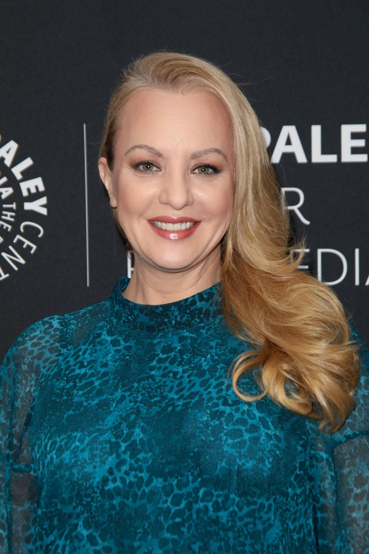 Celebrity 桌布 壁紙 — 一位微笑的金髮女性穿著青綠色豹紋上衣，在帶有白色文字的深色活動背景前擺姿勢。