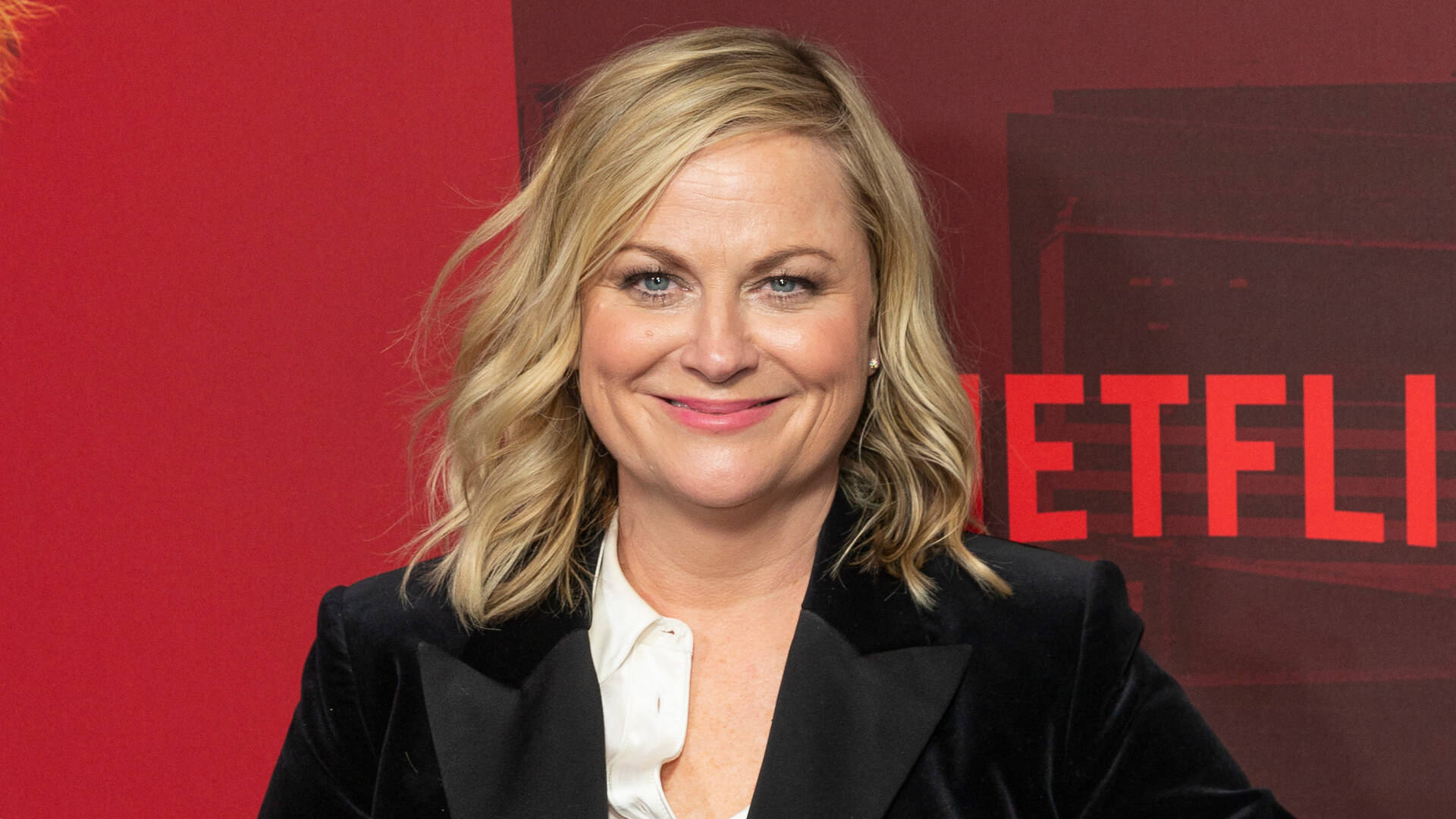 Netflix 桌布 壁紙 — 一位微笑的金髮女性穿著黑色西裝外套，站在活動中的紅色Netflix背景前。