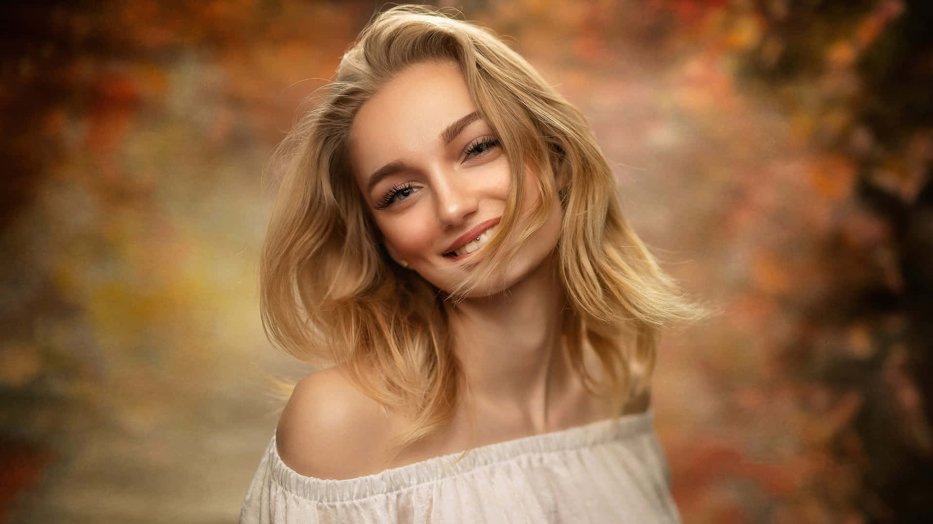 美女 桌布 壁紙 — 一位微笑的金髮女性身穿白色露肩上衣，站在溫暖的秋葉背景前。