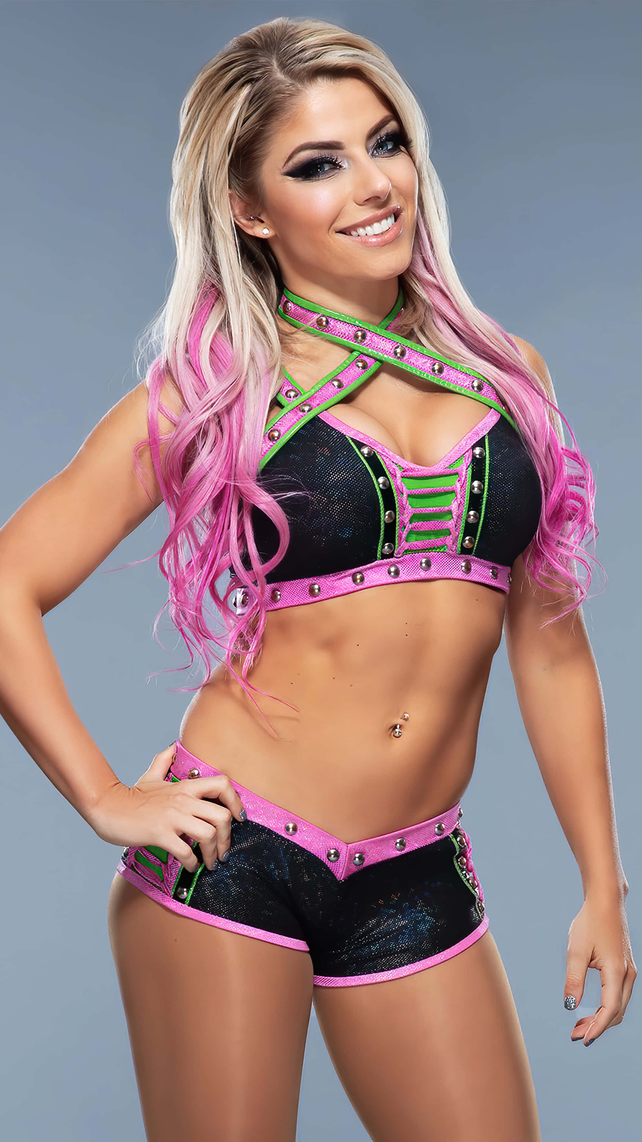WWE 桌布 壁紙 — 一位微笑的金髮女摔角手，帶有粉紅色接髮，穿著黑色和粉紅色鉚釘服裝，在灰色工作室背景前自信地擺姿勢。