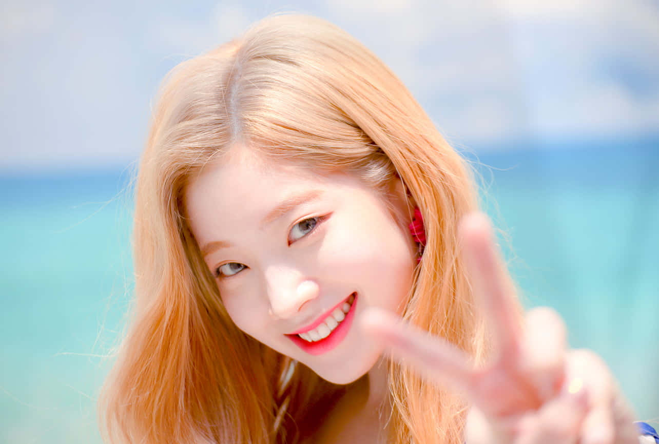 twice 桌布 壁紙 — 一位微笑的金髮年輕女性在明亮的藍綠色海洋背景下做出和平手勢。