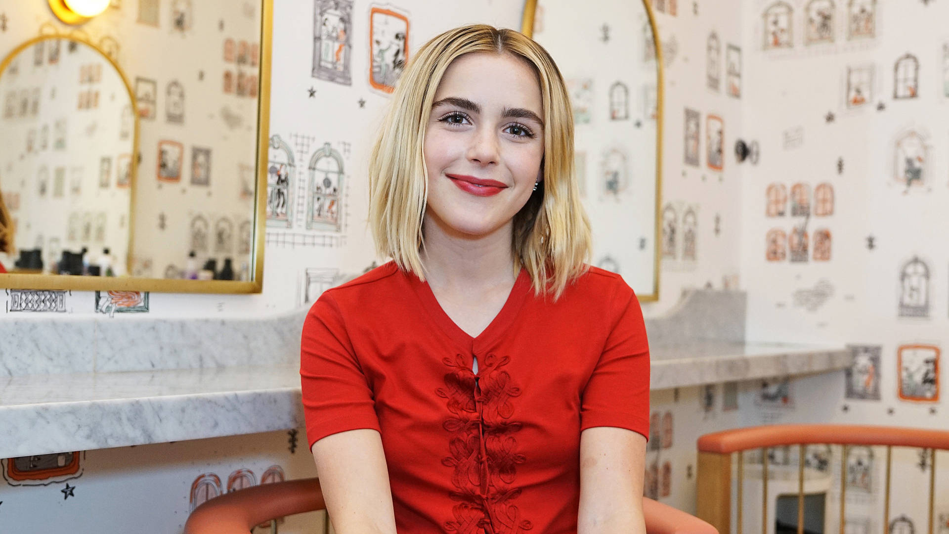 Kiernan Shipka 桌布 壁紙 — 一位微笑的金髮年輕女性穿著紅色上衣，背景是俏皮的插圖壁紙。
