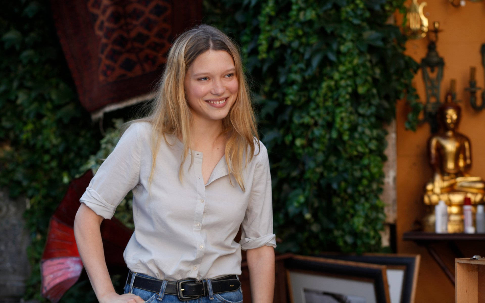 Lea Seydoux 桌布 壁紙 — 一位微笑的金髮年輕女性自信地站在一個充滿陽光、綠植和金色裝飾的房間裡。