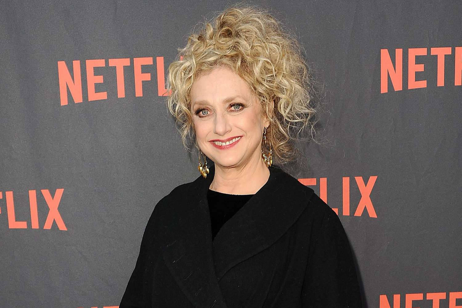 Actress HD 桌布 壁紙 — 一位微笑的金髮捲髮女性，身穿黑色外套，背景為灰色Netflix品牌背景。