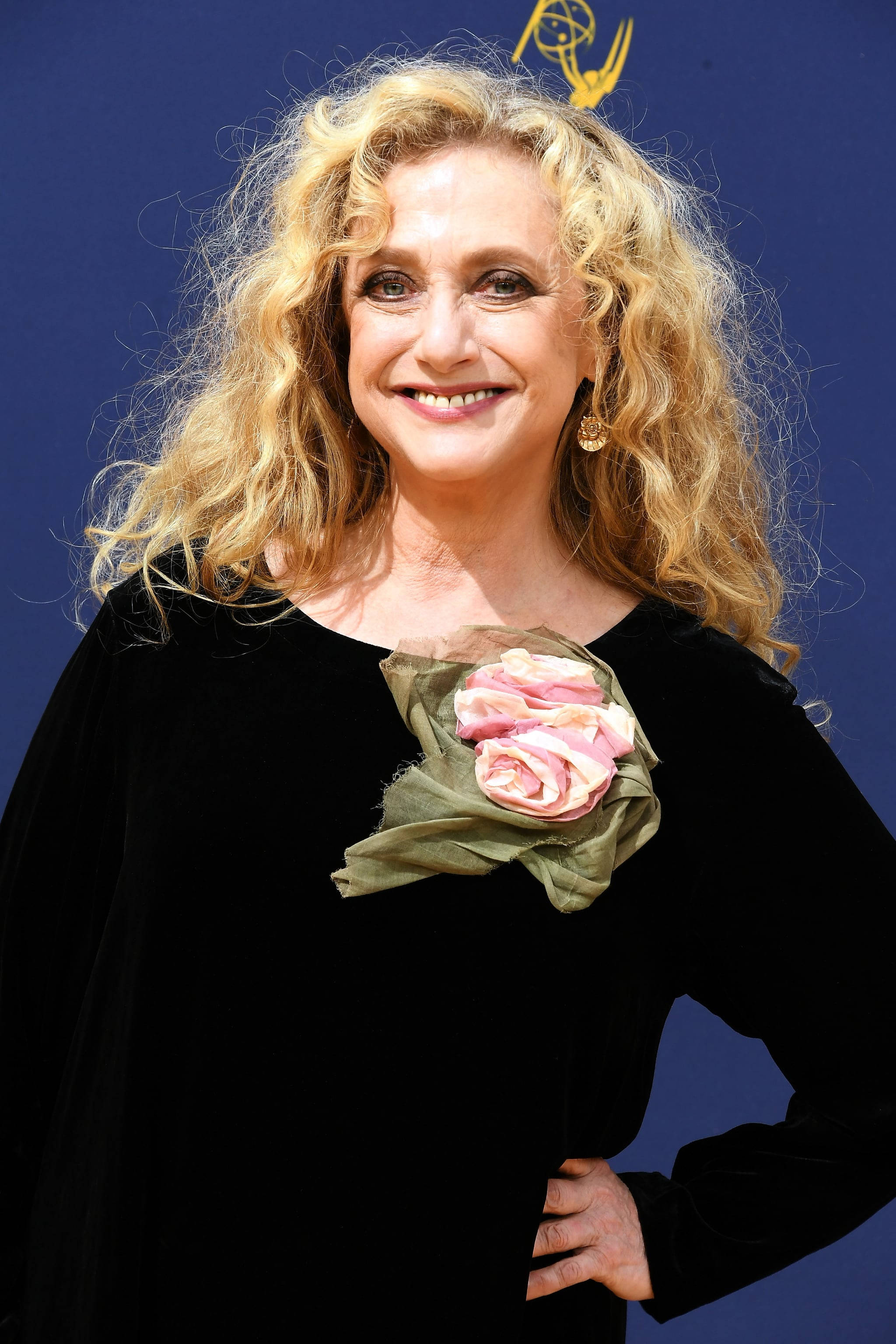 Actress HD 桌布 壁紙 — 一位微笑的金髮捲髮女性，身穿黑色天鵝絨連衣裙，飾有粉紅色玫瑰胸花，背景為藍色。
