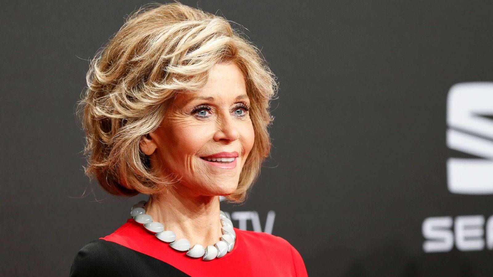 Celebrity 桌布 壁紙 — 一位微笑的金髮波浪長髮女性，身穿紅黑上衣，戴著白色珠鍊，背景為深色，帶有部分白色文字。